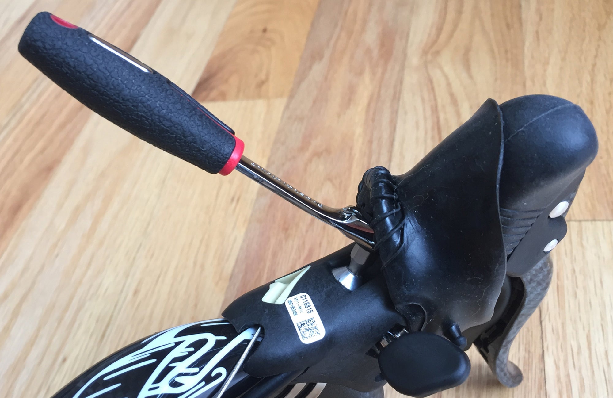 A close-up of a bike handlebar with a black rubber brake lever, showing the Prestacycle Prestaratchet Pro Mini Ratchet Bicycle Tool wedged under the hood, against a light wood floor background.