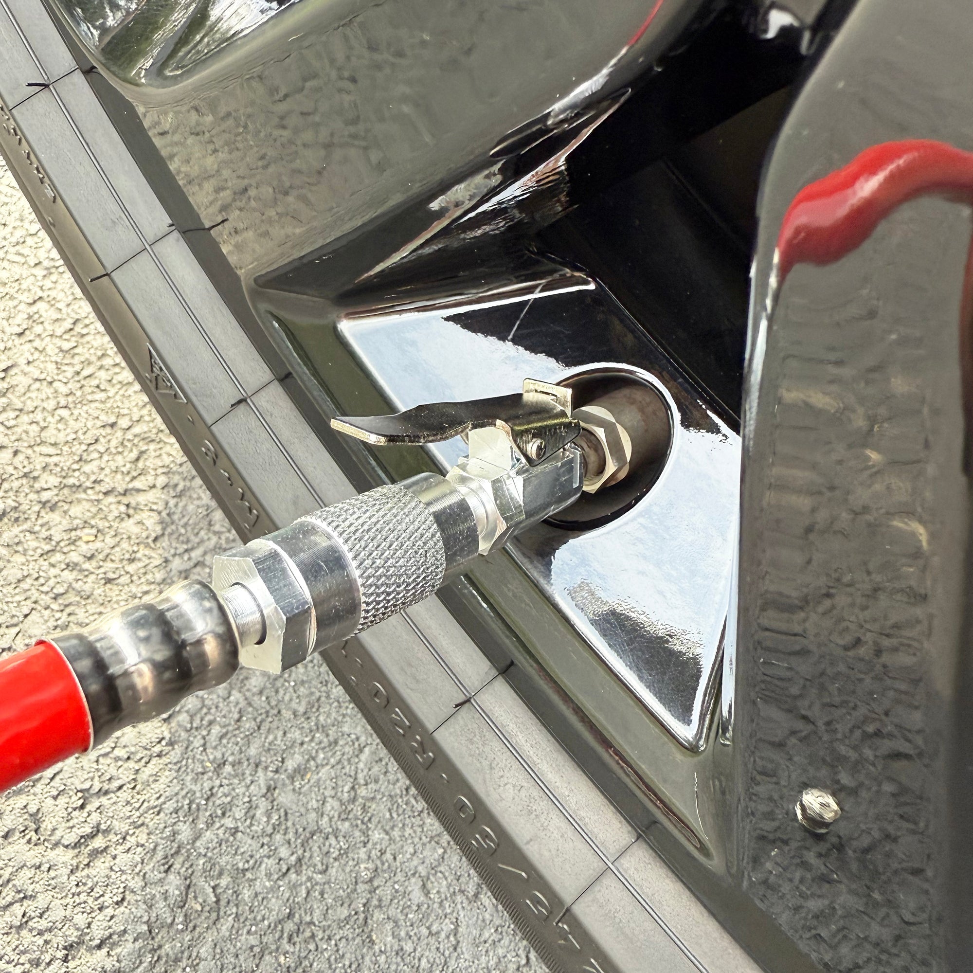 A close-up of the Prestacycle Prestaflator Bicycle Tire Inflator attached to a tire valve, inflating a car tire. The shiny black car and textured road surface are visible in the background. Compatible with Presta and Schrader valves.