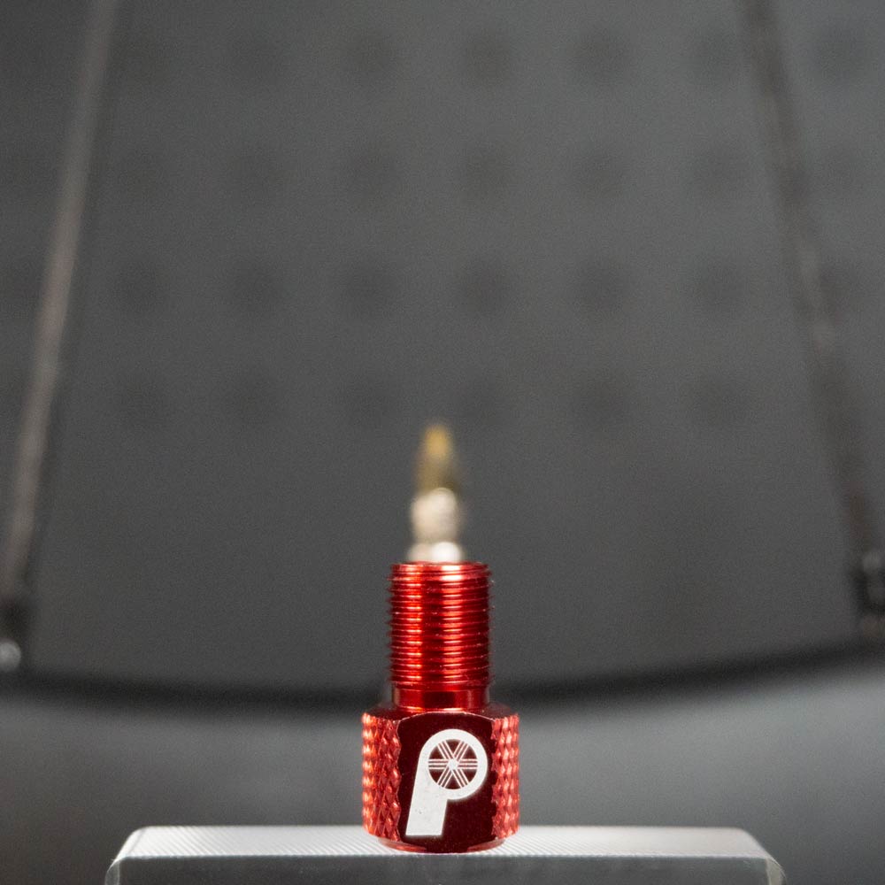 The Prestacycle Prestadapter TUBELESS, a red lightweight alloy Presta adapter and valve core remover with a white wheel logo, stands upright on a silver surface against a blurred, dark background.