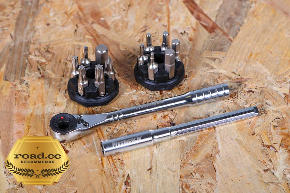 The Prestacycle Pro T-Handle Kit by Prestacycle, featuring a T-handle ratchet, extension bar, two circular bit holders with assorted 1/4 bits, is displayed on wood. A gold road.cc recommended badge appears in the bottom left corner.