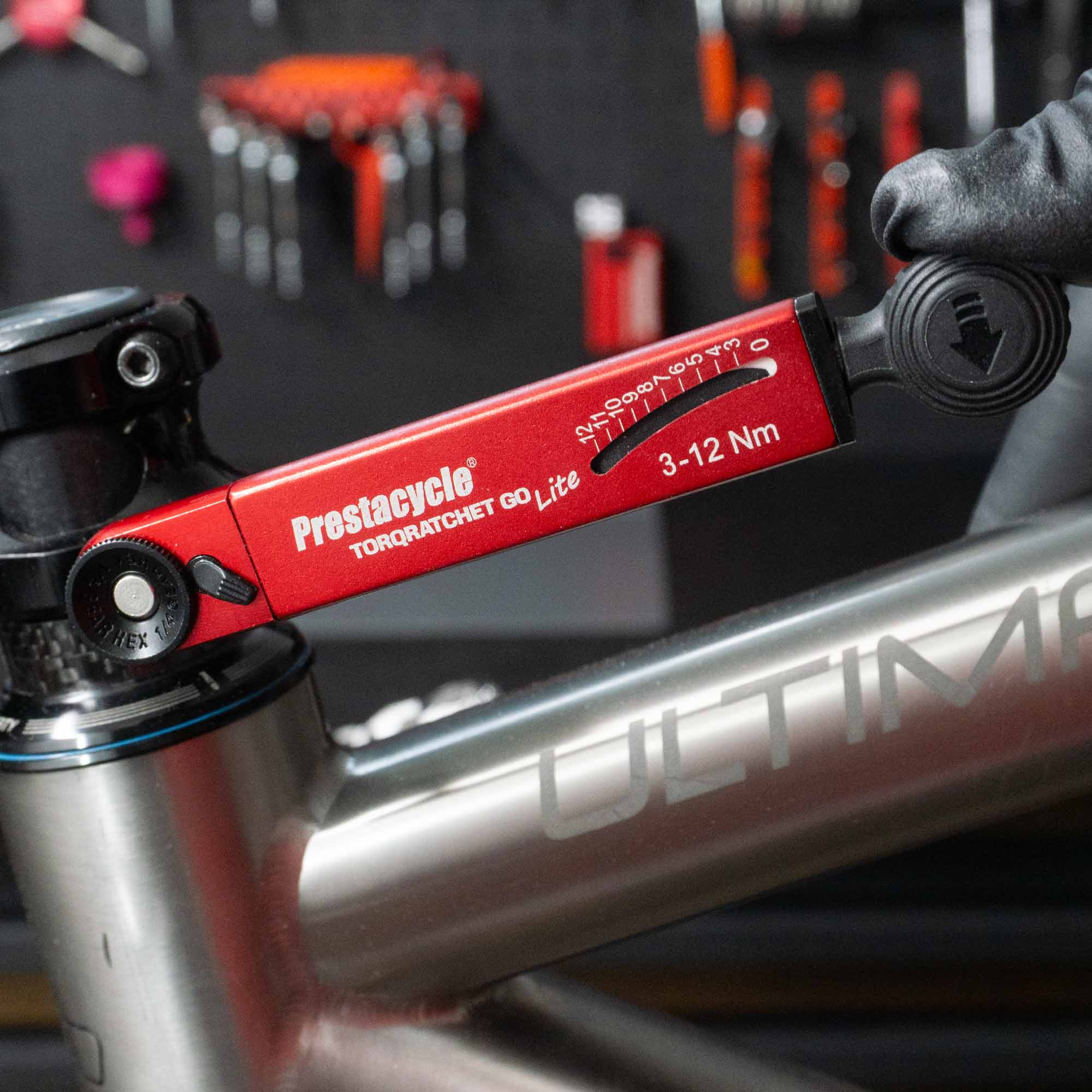 A hand uses a red Prestacycle TorqRatchet GO Lite - Portable 3~12Nm Torque Multi-tool Ratchet to adjust a bolt on a silver ULTIMA bicycle frame, with bike tools hanging on a wall in the blurred background.
