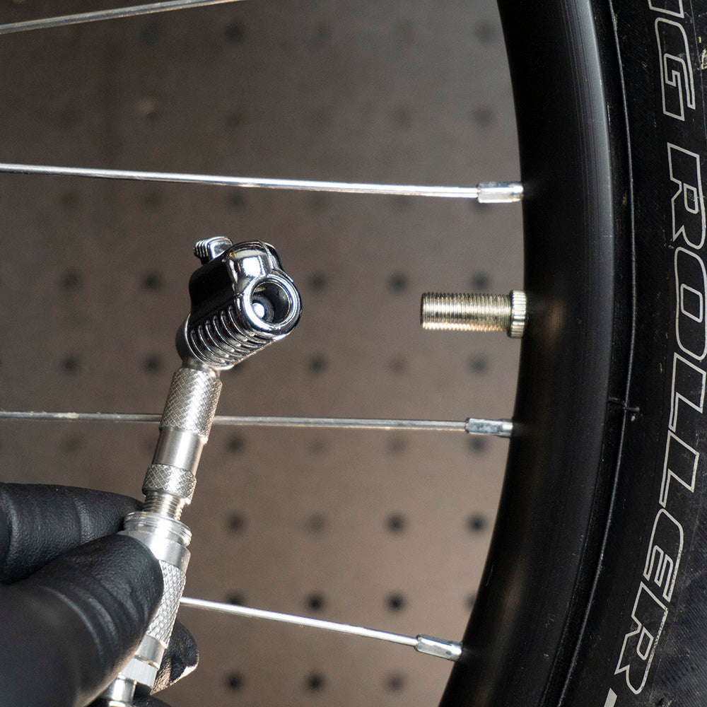 A gloved hand holds the Prestacycle Pro Schrader Head by Prestacycle, featuring an auto-lock mechanism, near a Schrader valve on a bicycle wheel, ready to check tire pressure.