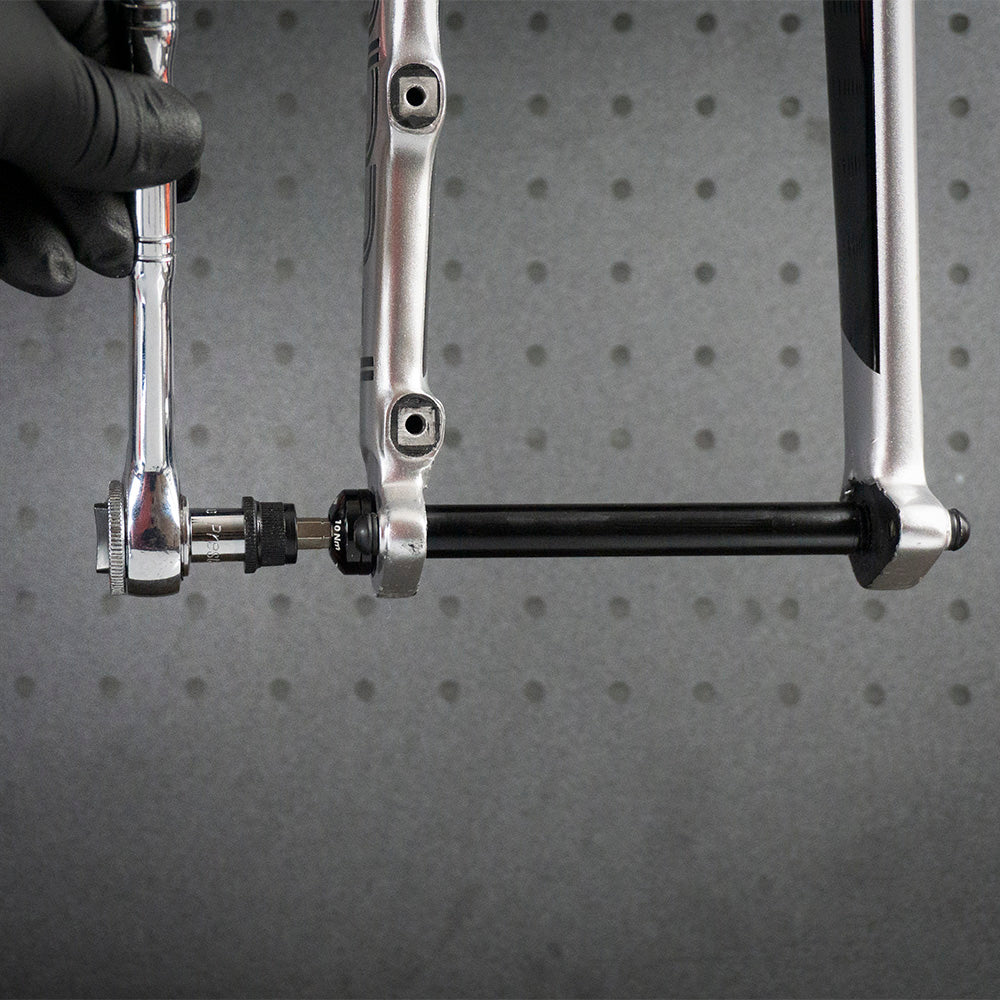 A gloved hand uses a Prestacycle 1/4” Square to Hex Bits Quick Release Adapter to tighten a bolt on a bicycle axle, with part of the bike frame and the adapter visible against a gray, perforated background.
