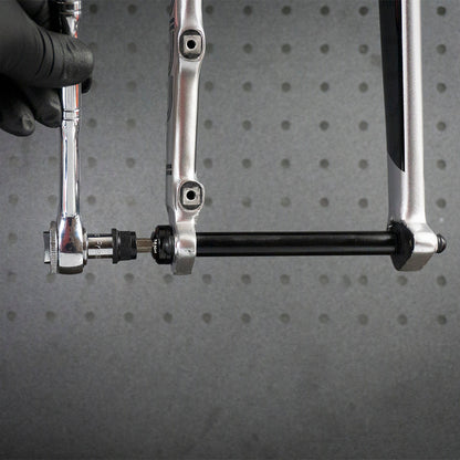 A gloved hand uses a Prestacycle 1/4” Square to Hex Bits Quick Release Adapter to tighten a bolt on a bicycle axle, with part of the bike frame and the adapter visible against a gray, perforated background.