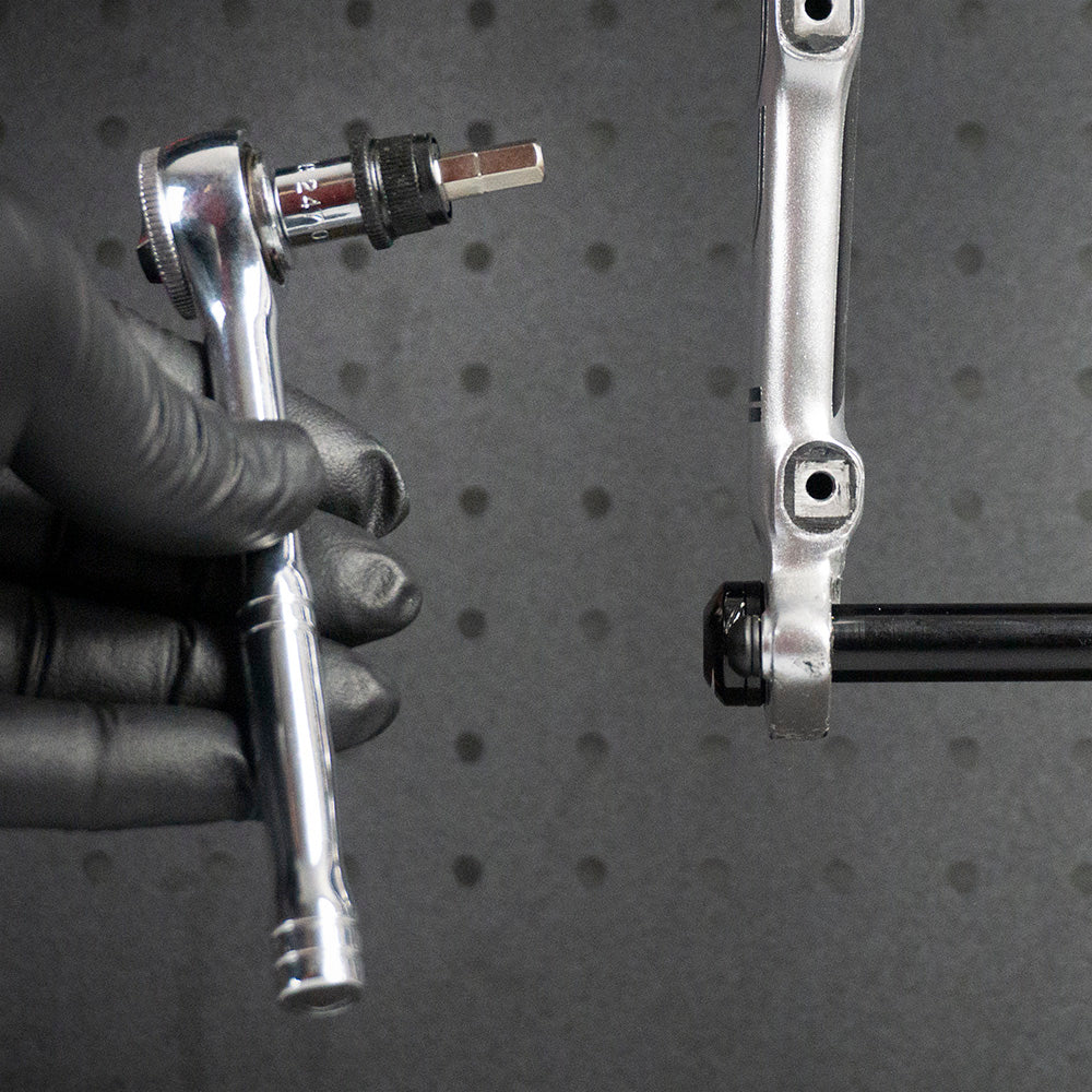 A gloved hand uses the Prestacycle 1/4” Square to Hex Bits Quick Release Adapter near a silver bike crank arm, ready to adjust a bolt. The dark, perforated background is ideal for 1/4″ socket-type torque wrenches.