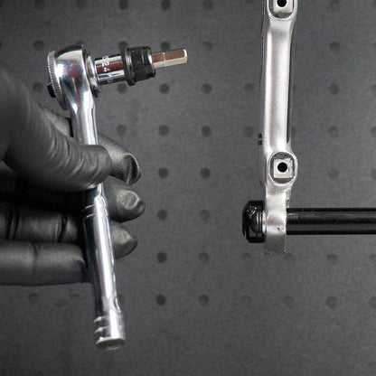 A gloved hand uses the Prestacycle 1/4” Square to Hex Bits Quick Release Adapter near a silver bike crank arm, ready to adjust a bolt. The dark, perforated background is ideal for 1/4″ socket-type torque wrenches.