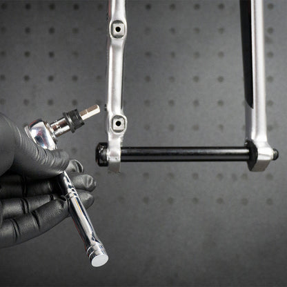 A gloved hand holds the Prestacycle 1/4” Square to Hex Bits Quick Release Adapter near a bicycle frame dropout, preparing to install or tighten a thru-axle. A black pegboard is visible in the background.