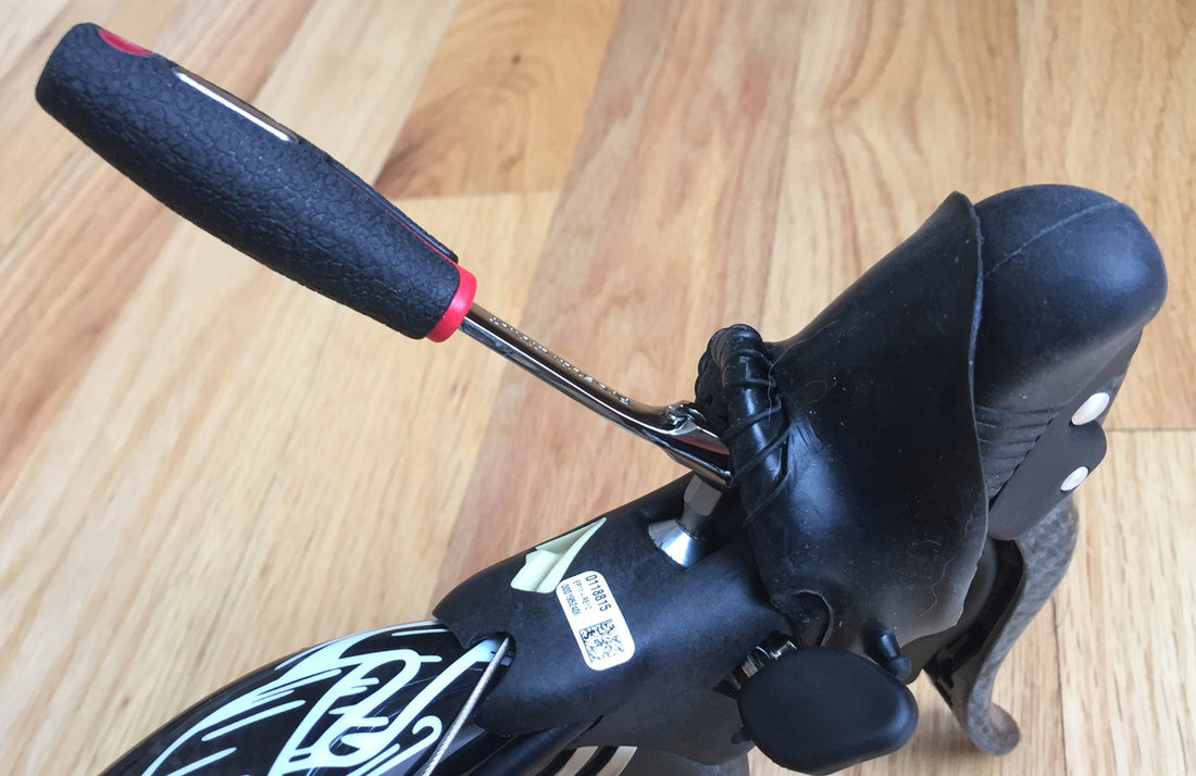 A close-up of a bike handlebar with a black rubber brake lever, showing the Prestacycle Prestaratchet Pro Mini Ratchet Bicycle Tool wedged under the hood, against a light wood floor background.