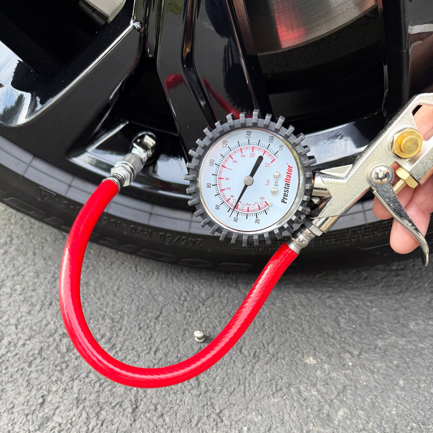 A close-up of a Prestacycle Prestaflator Bicycle Tire Inflator with red hose connected to a tire valve, showing the pressure reading. The Presta &amp; Schrader air compressor tool is attached to the visible wheel on gray pavement.