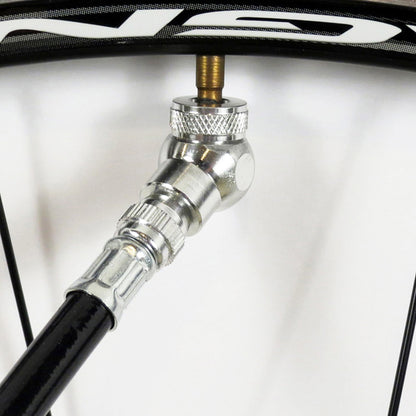 A close-up of a bicycle tubeless tire valve being inflated with the Prestacycle Angle Mini Presta Head pump nozzle attached to the valve stem. Black wheel spokes are visible in the background.