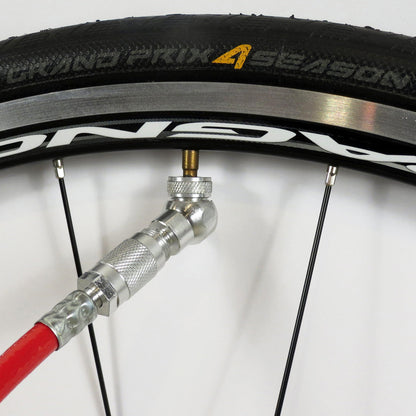 A close-up shows a Prestacycle Angle Mini Presta Head pump nozzle attached to the valve of a Grand Prix 4 Season tubeless tire, with the wheel rim and spokes also visible.