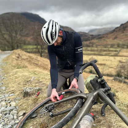 A cyclist in full gear uses the Prestacycle Prestaflator GO Portable Digital Rechargeable Preset Bicycle Tire Inflator to repair a front wheel on a rocky path, with rolling hills and a cloudy sky in the background.