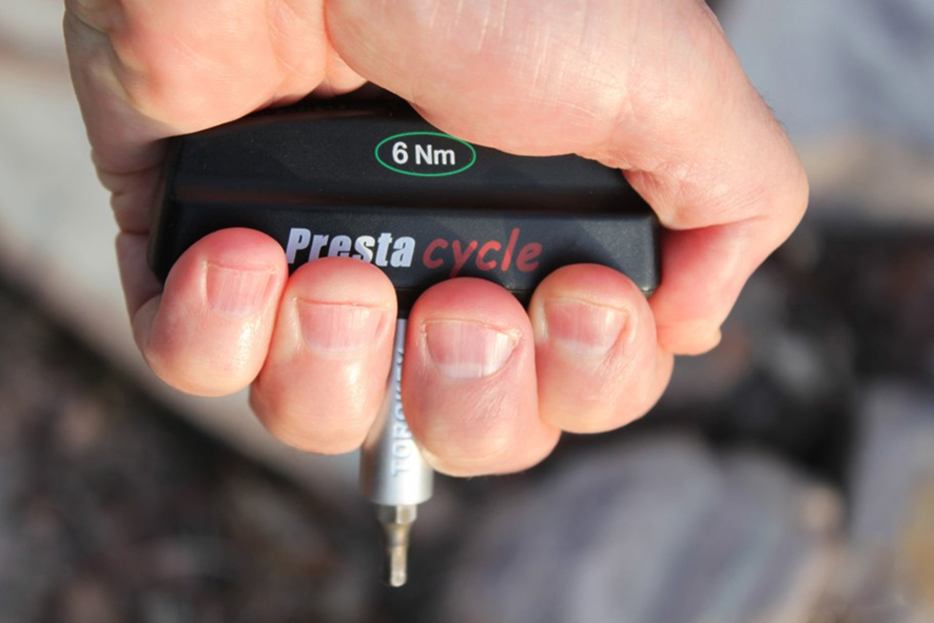 A close-up shows a hand gripping the Prestacycle Pro TorqKeys - 6Nm T-Handle Torque-Limiting Bits Tool, featuring the 6 Nm label and tool tip. The black ergonomic handle stands out against a blurred natural outdoor background.