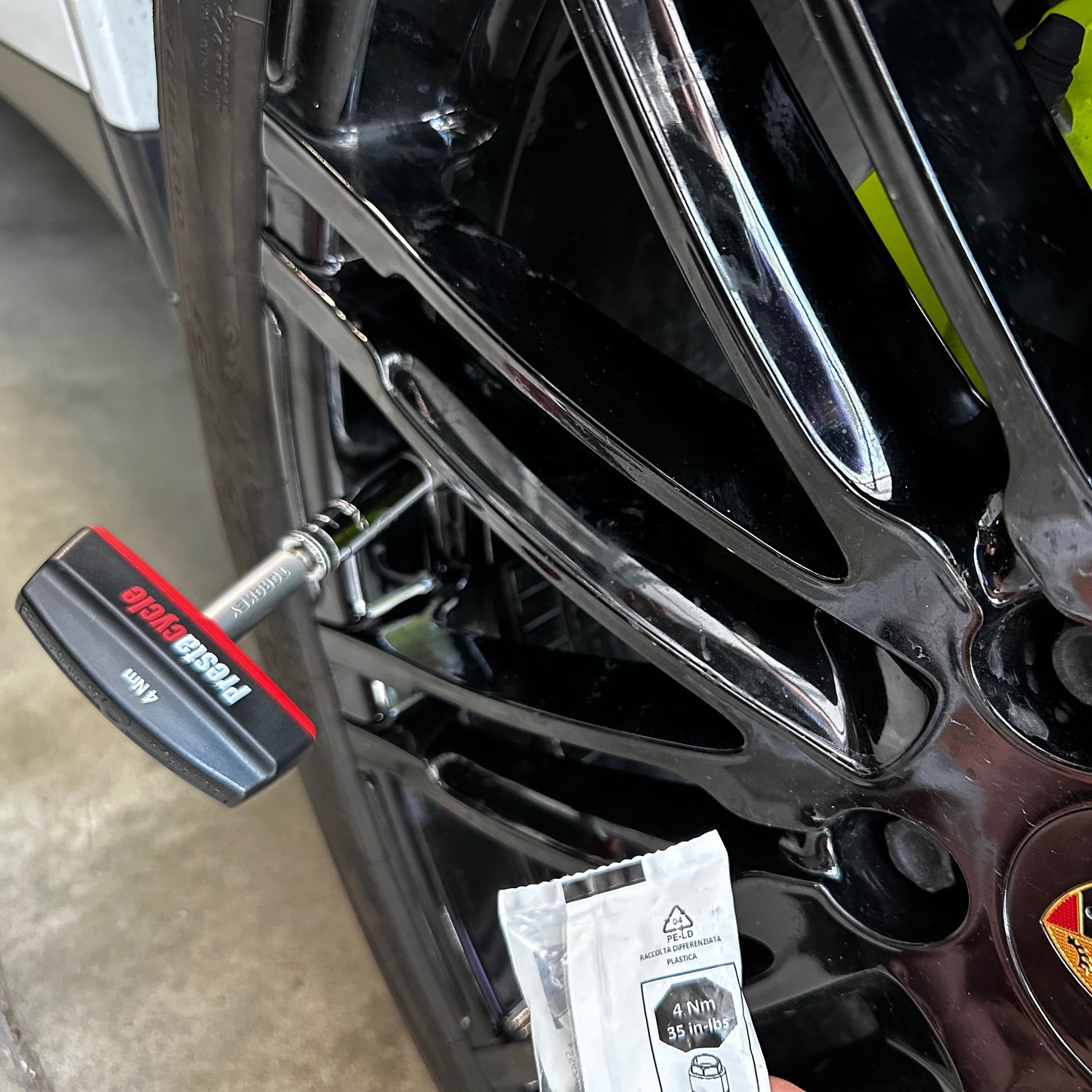 A close-up shows a black car wheel with a tire pressure gauge on the valve. A hand holds the Prestacycle Pro TorqKeys 4Nm T-Handle Torque-Limiting Bits Tool near the wheel. The car is parked indoors.