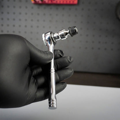 A hand in a black glove holds the Prestacycle Bits Swivel QR Torque Wrench Adapter by Prestacycle, a shiny metal tool with socket attachment, against a dark perforated background.