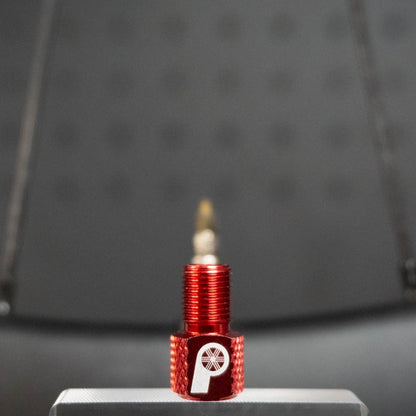 The Prestacycle Prestadapter TUBELESS, a red lightweight alloy Presta adapter and valve core remover with a white wheel logo, stands upright on a silver surface against a blurred, dark background.