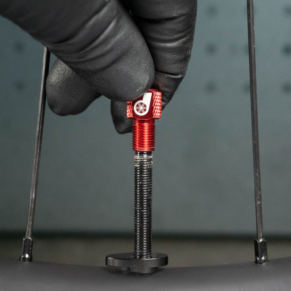 A gloved hand adjusts a Prestacycle Prestadapter TUBELESS—Presta adapter and valve core remover—in red anodized alloy, attached to a threaded rod. Two black spokes of the bicycle wheel are visible on either side.