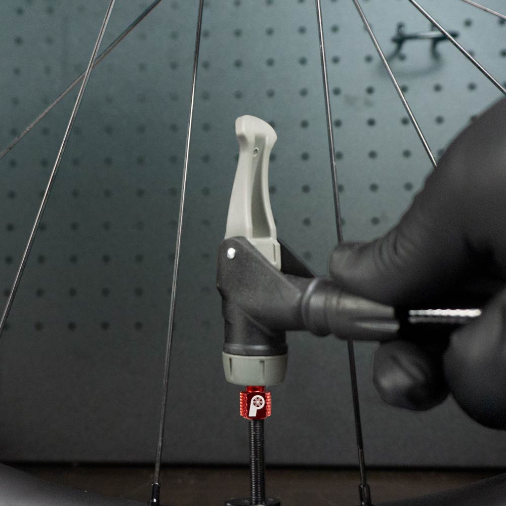 A gloved hand inflates a bicycle tire using the Prestacycle Prestadapter TUBELESS – a Presta adapter and valve core remover – attaching it to a red Presta valve on a lightweight alloy wheel with visible metal spokes in the background.