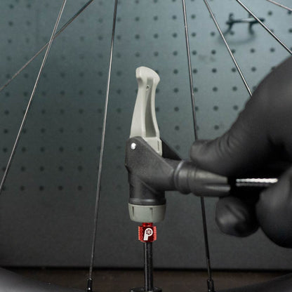 A gloved hand inflates a bicycle tire using the Prestacycle Prestadapter TUBELESS – a Presta adapter and valve core remover – attaching it to a red Presta valve on a lightweight alloy wheel with visible metal spokes in the background.