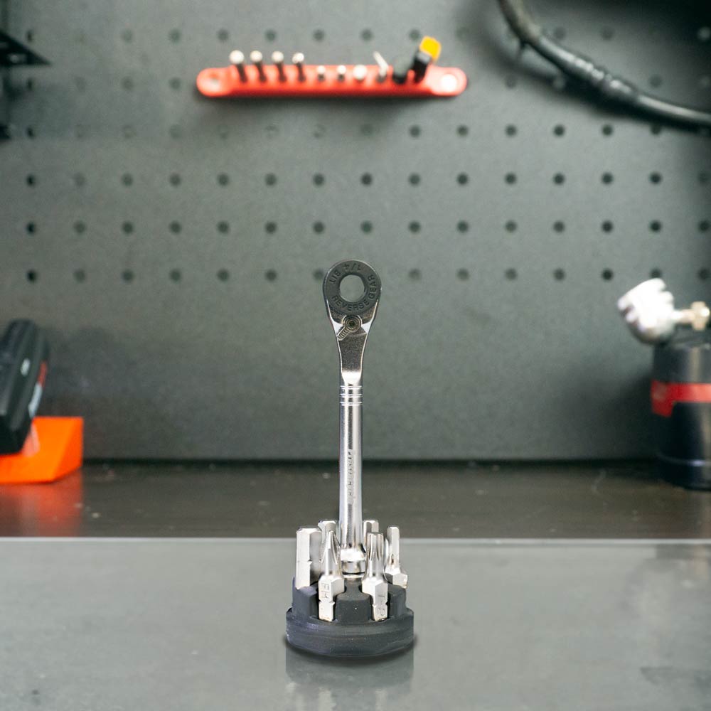 The Prestacycle Prestaratchet Micro – Kit with 6 S2 bits and a round holder stand is displayed upright on a workbench, with the bits arranged in the circular holder and tools visible on the pegboard in the background.