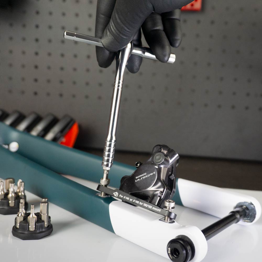 A gloved hand uses the Prestacycle Pro T-Handle Kit to adjust a bicycle disc brake caliper, with 1/4 inch bits and other Prestacycle tools visible on the workbench in the background.