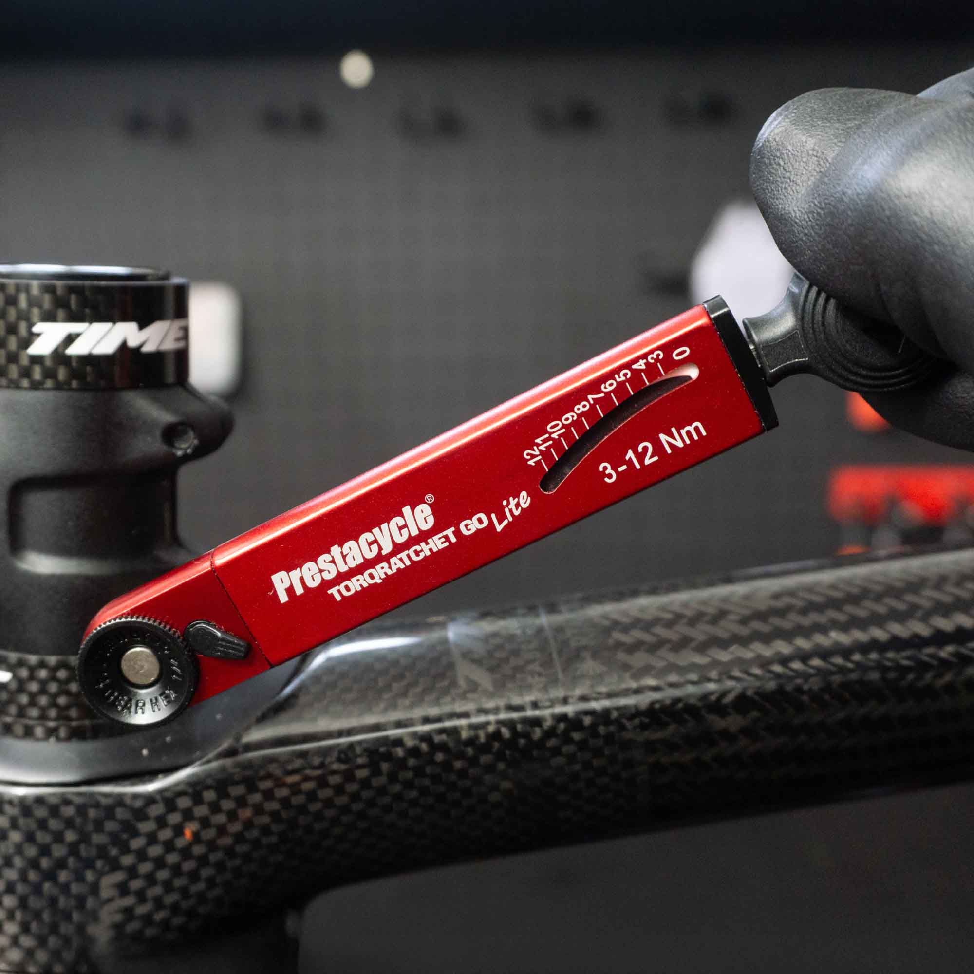 A gloved hand uses the Prestacycle TorqRatchet GO Lite—an adjustable 3–12Nm portable torque multi-tool ratchet—to adjust a bolt on a black bicycle frame.