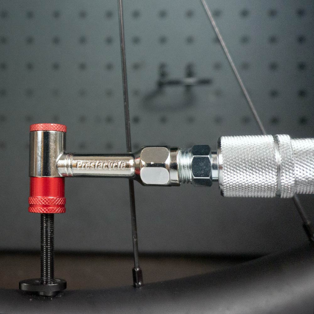 A close-up of the Prestacycle Prestaflator Mini Pro, a Presta/Schrader push-to-inflate bicycle inflation tool, attached to a cycling valve with its metal and red valve tool in use, set against a blurred workshop background.