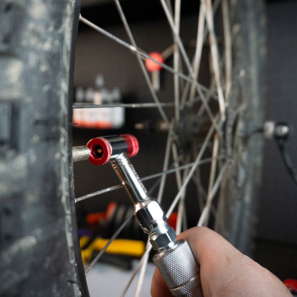 A close-up of a hand using the Prestacycle Prestaflator Mini Pro Bicycle Inflation Tool to check air pressure on a bike tire, highlighting its silver body, red fitting, cycling valve, and the bike wheel’s spokes and tread.