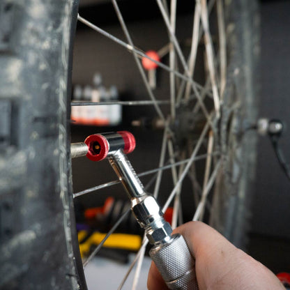 A close-up of a hand using the Prestacycle Prestaflator Mini Pro Bicycle Inflation Tool to check air pressure on a bike tire, highlighting its silver body, red fitting, cycling valve, and the bike wheel’s spokes and tread.