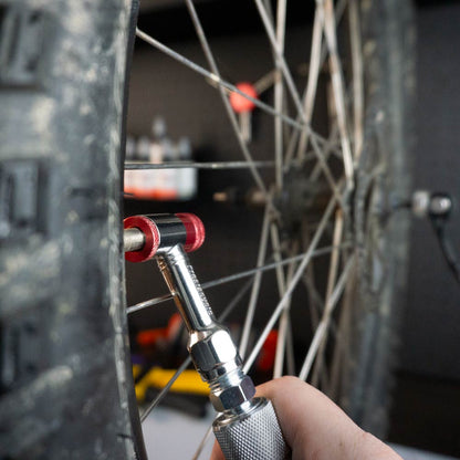 A close-up shows a hand using the Prestacycle Prestaflator Mini Pro – Presta / Schrader push-to-inflate Bicycle Inflation Tool to check air pressure, with the bike’s wheel, spokes, and valve clearly visible.