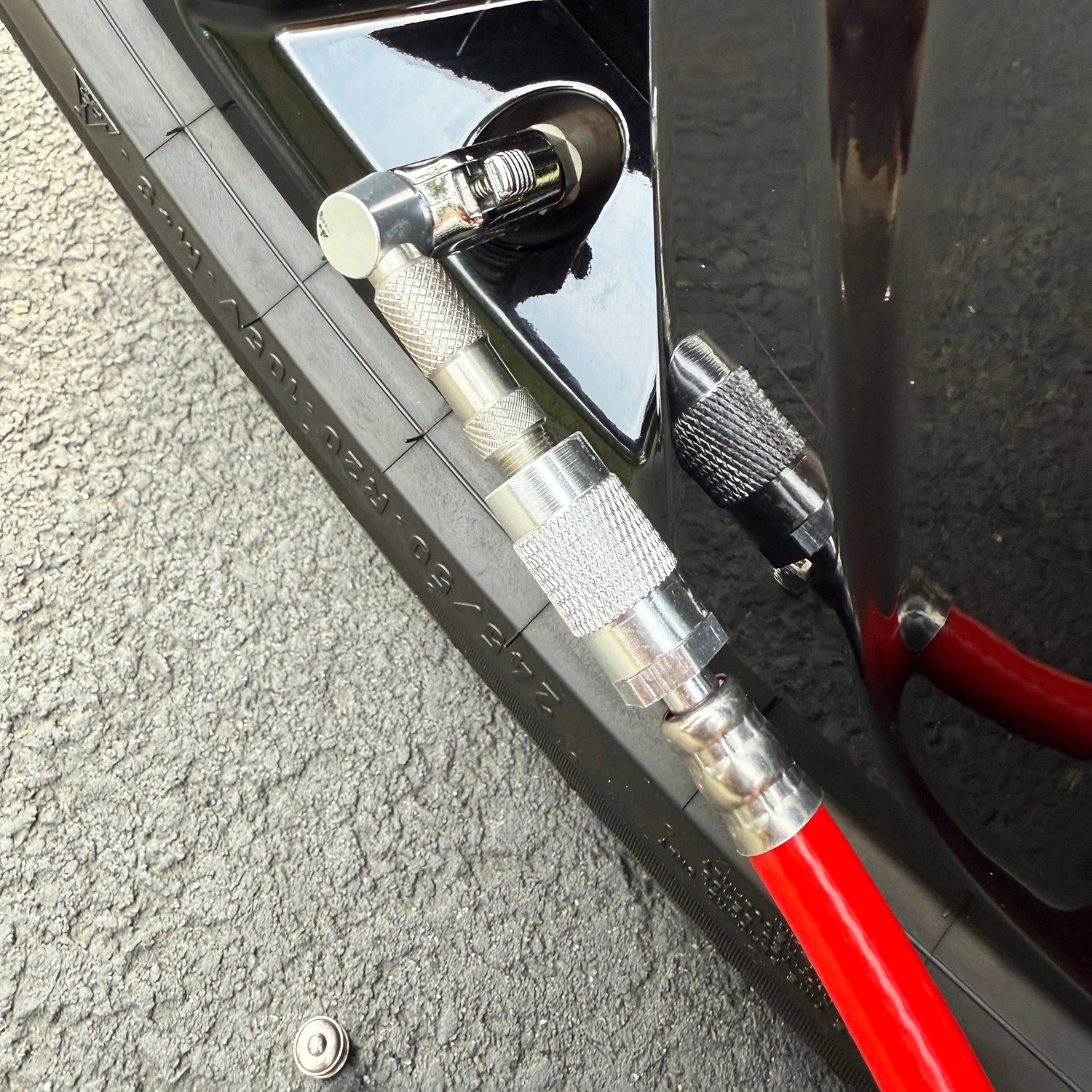 A close-up shows a tire being inflated on asphalt with a red air hose featuring the Prestacycle Pro Schrader Head by Prestacycle attached to a cars valve stem, highlighting the tire tread and ensuring accurate pressure.