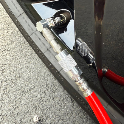 A close-up shows a tire being inflated on asphalt with a red air hose featuring the Prestacycle Pro Schrader Head by Prestacycle attached to a cars valve stem, highlighting the tire tread and ensuring accurate pressure.