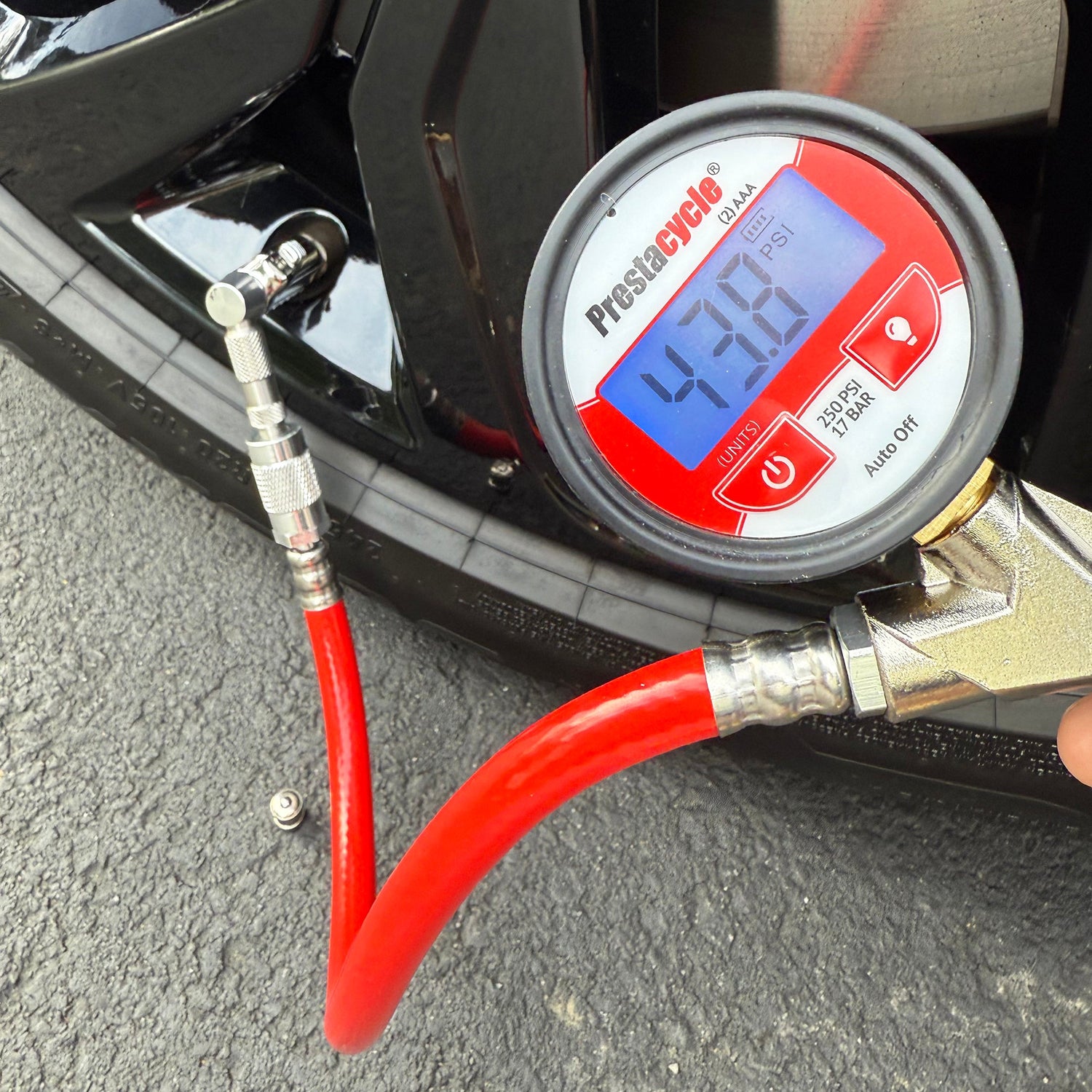 The Prestacycle Pro Schrader Head digital tire pressure gauge by Prestacycle shows 43.8 PSI while attached to a car tire valve with its red hose and auto-lock, with the tire and part of the wheel in view.