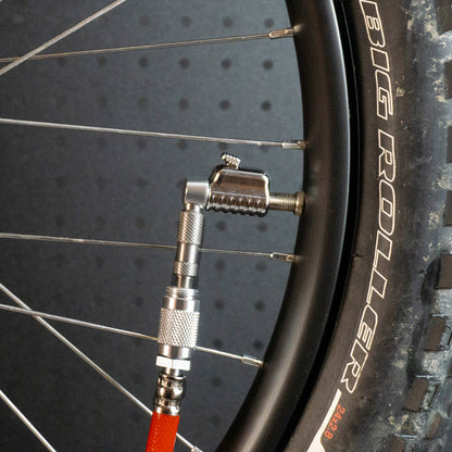 A close-up shows a bicycle tire being inflated with a metal air pump using the Prestacycle Pro Schrader Head by Prestacycle, securely attached to the valve stem on the wheel rim.