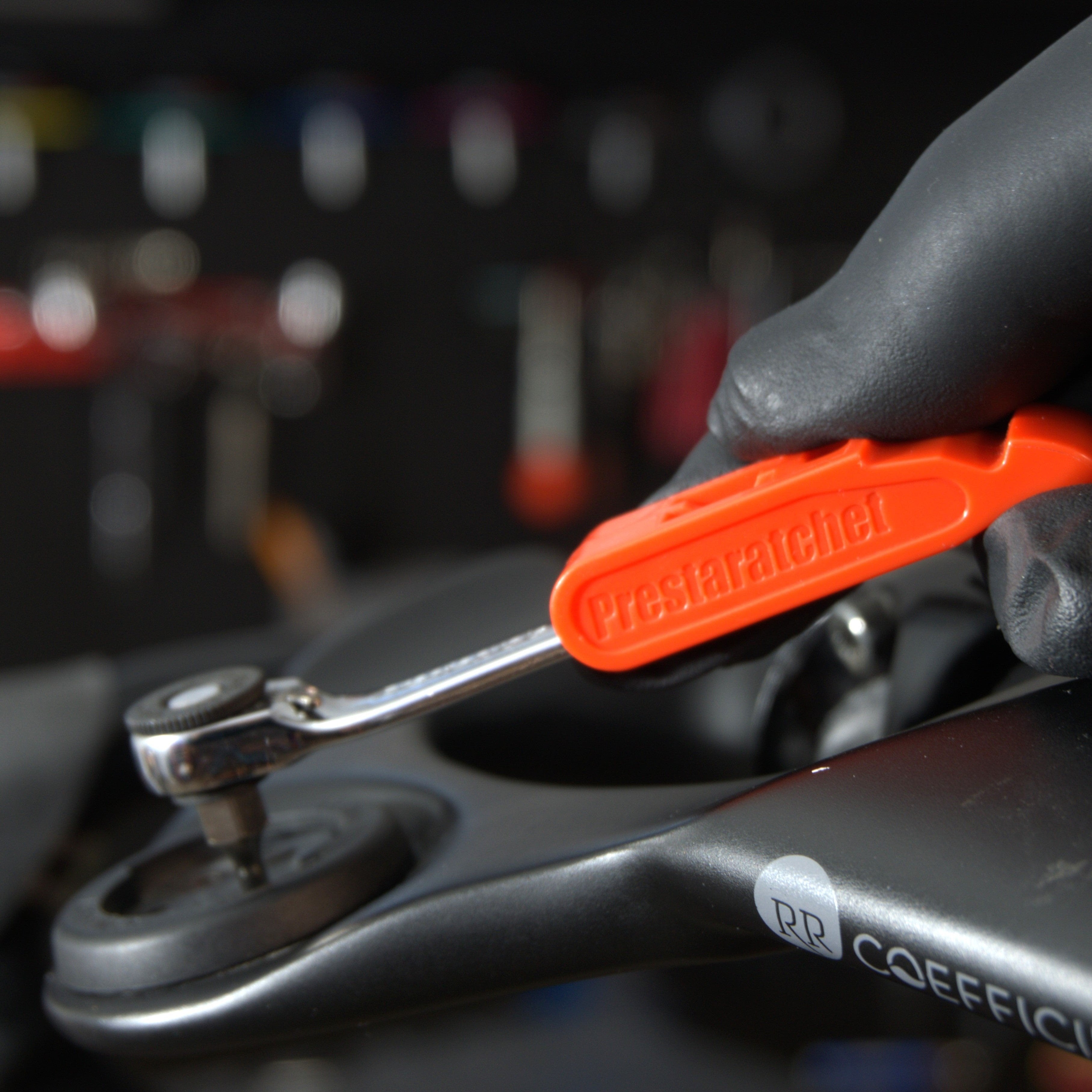 A gloved hand uses the Prestacycle Prestaratchet GO Multi-tool Kit to tighten a bolt on a dark-colored bike frame, with various tools blurred in the background.