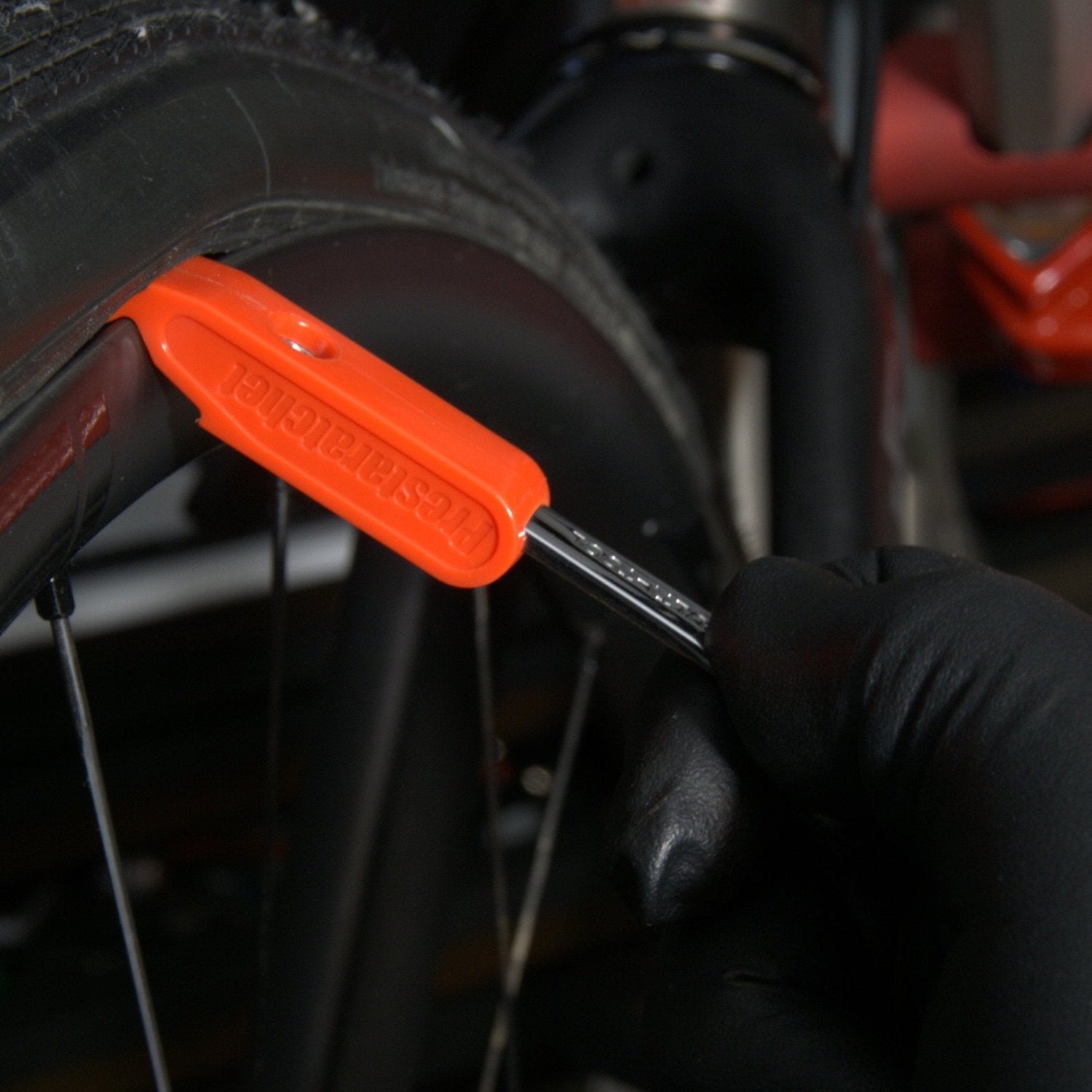 A gloved hand uses an orange tire lever to remove a bicycle tire, while the Prestacycle Prestaratchet GO 5-in-1 Multi-tool Kit is ready nearby for maintenance.